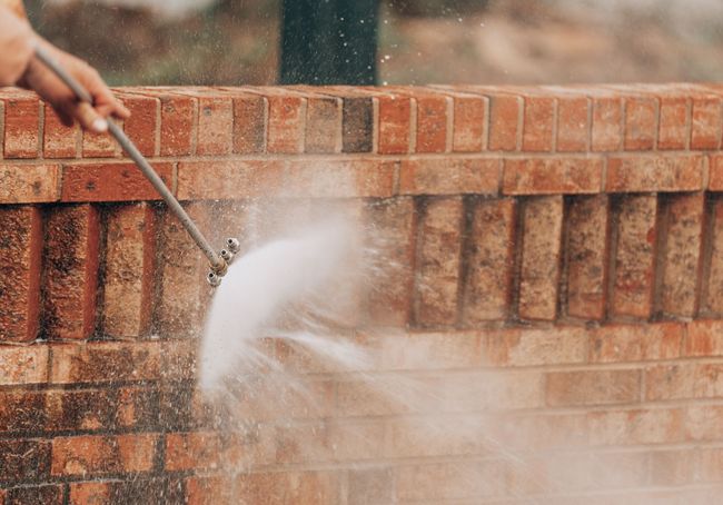 Person using a pressure washer to clean a brick wall, spraying water.