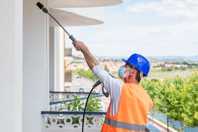 Man in safety gear spraying insecticide on building exterior.