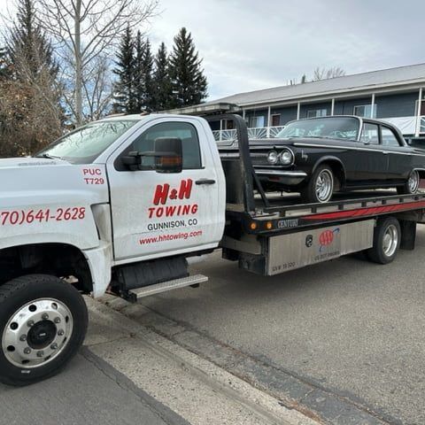 Tow truck with black classic car on its flatbed; in a town.