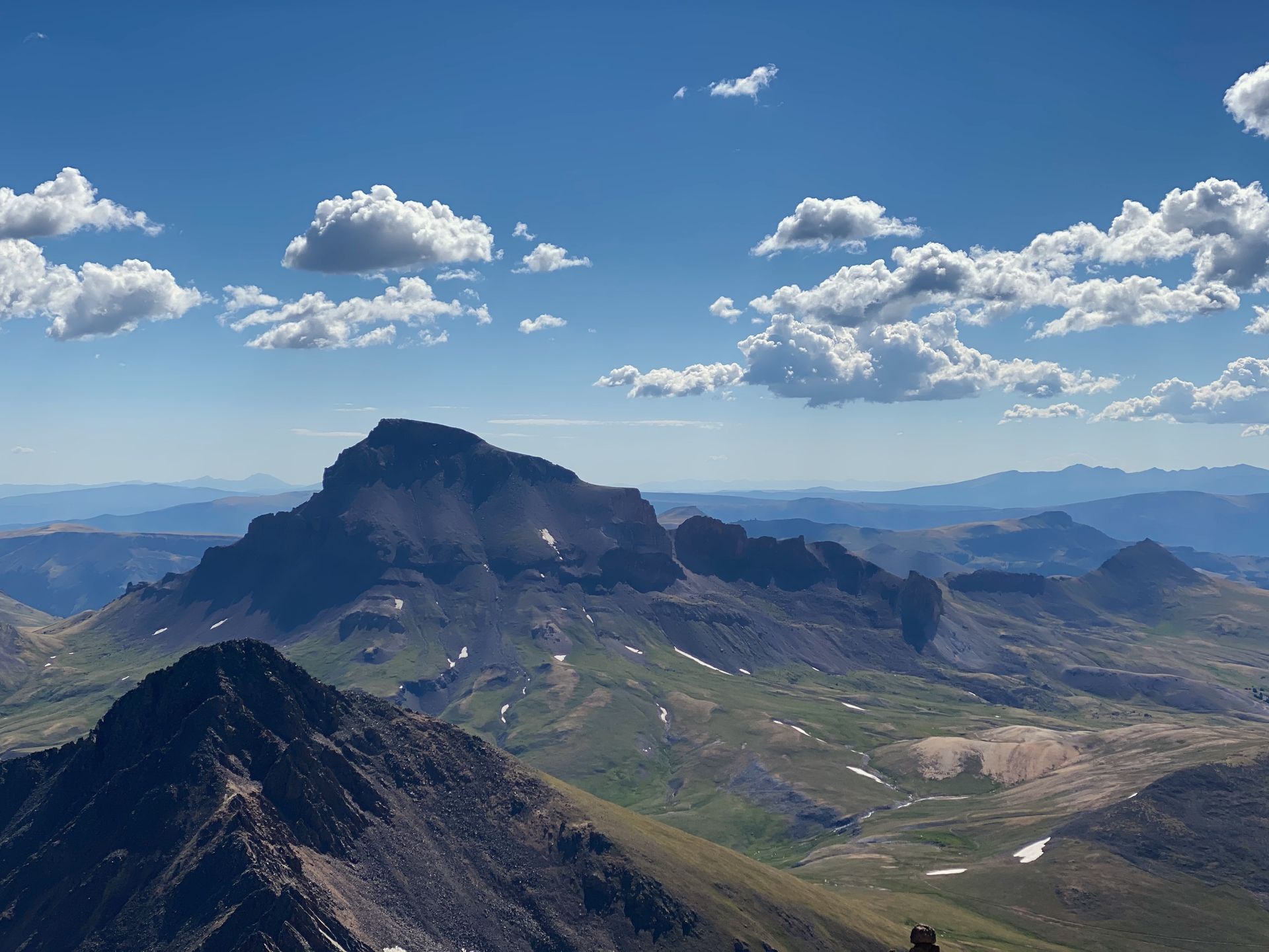 Mountain landscape under a blue sky with fluffy white clouds; green slopes, peaks, and valleys in view.