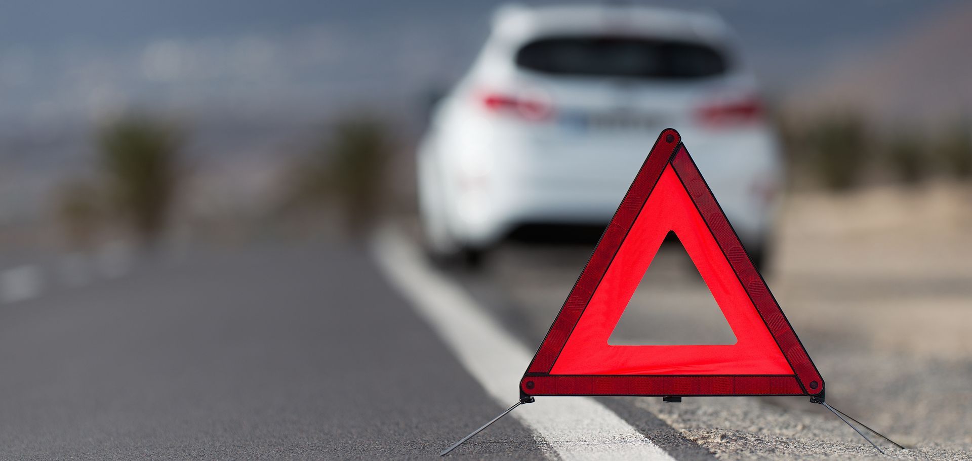 Red warning triangle on roadside with a white car in the blurred background.