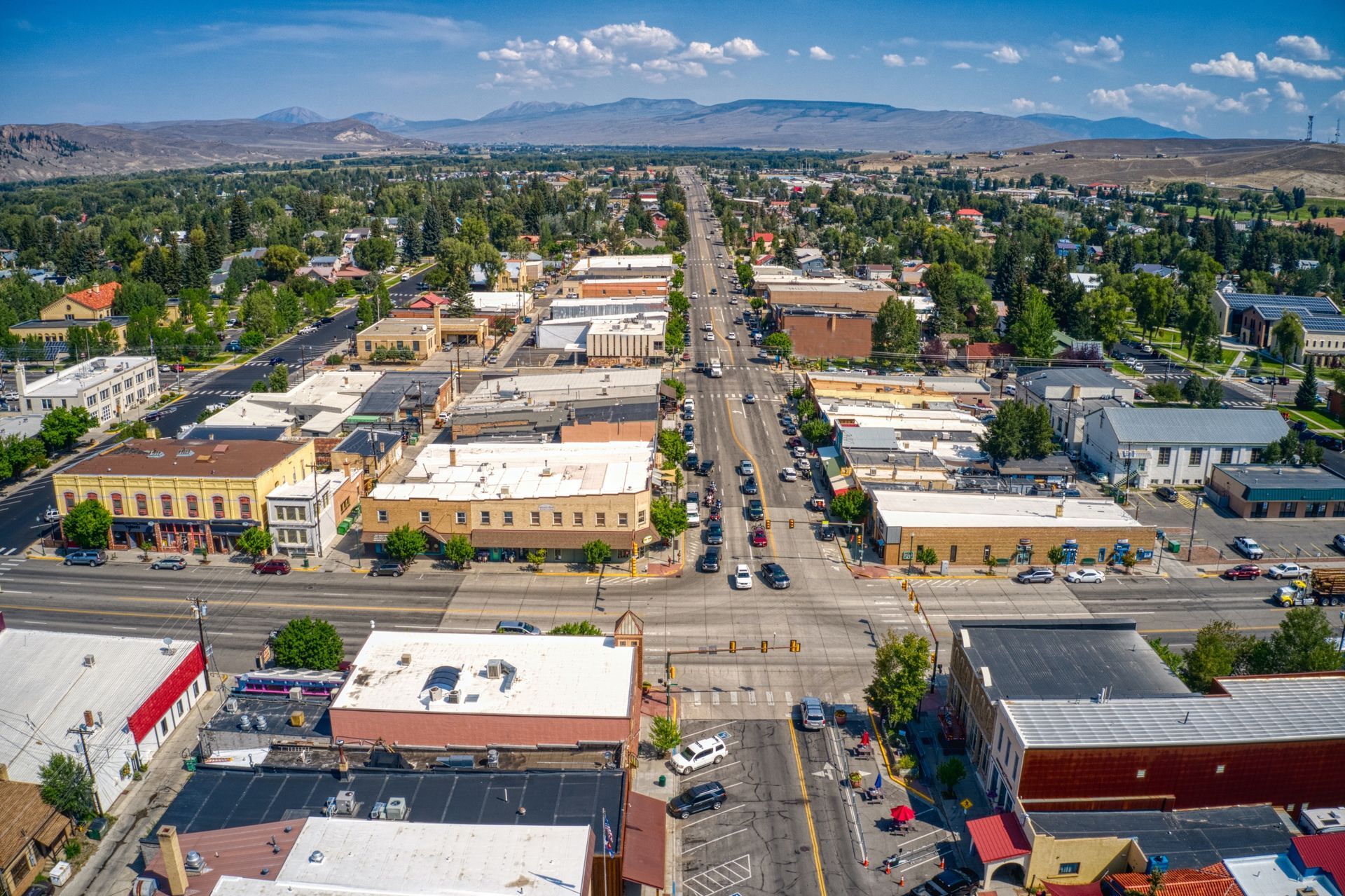 Aerial view of a town with buildings lining a road, mountains in the distance, and cars on the road.