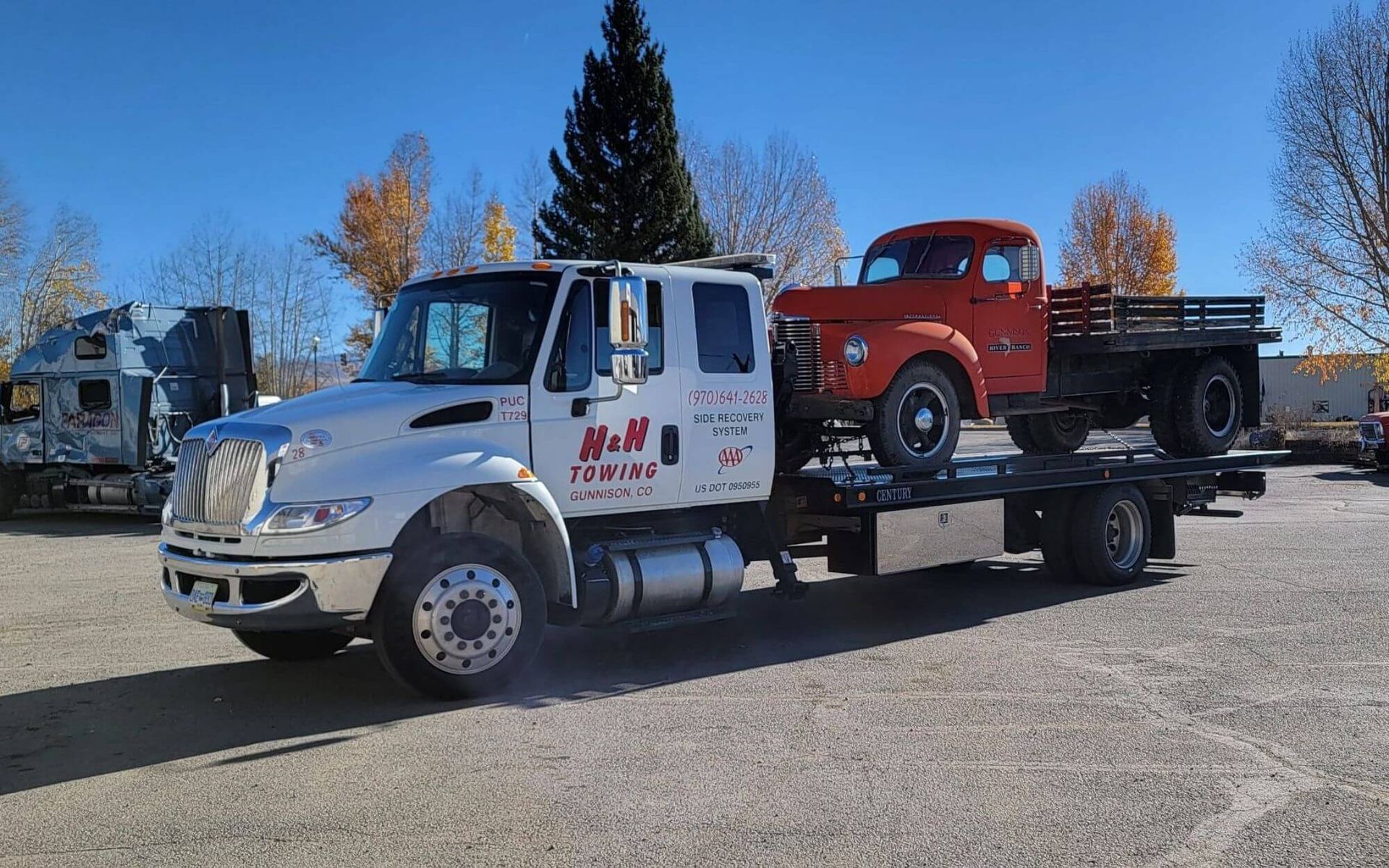White tow truck carrying an orange vintage flatbed truck on a sunny day.