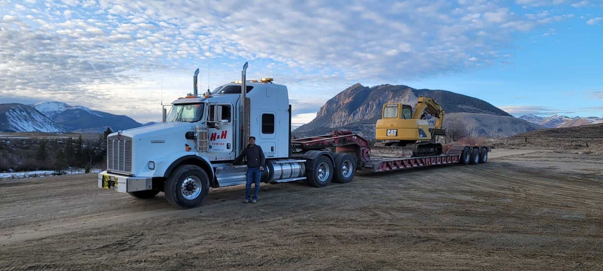 White semi-truck hauling yellow excavator on trailer in dirt field with mountains in background.