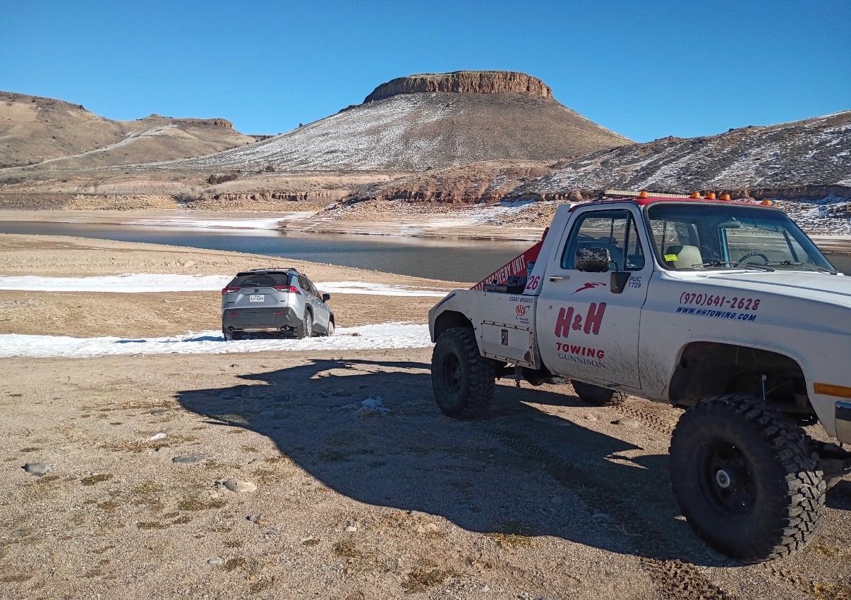 Tow truck pulling a gray SUV from near a lake, in a desert landscape with a mountain in the background.
