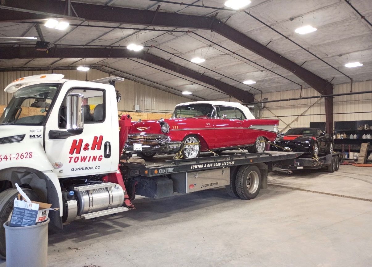 A red classic car on a tow truck inside a garage. Another car follows behind. 