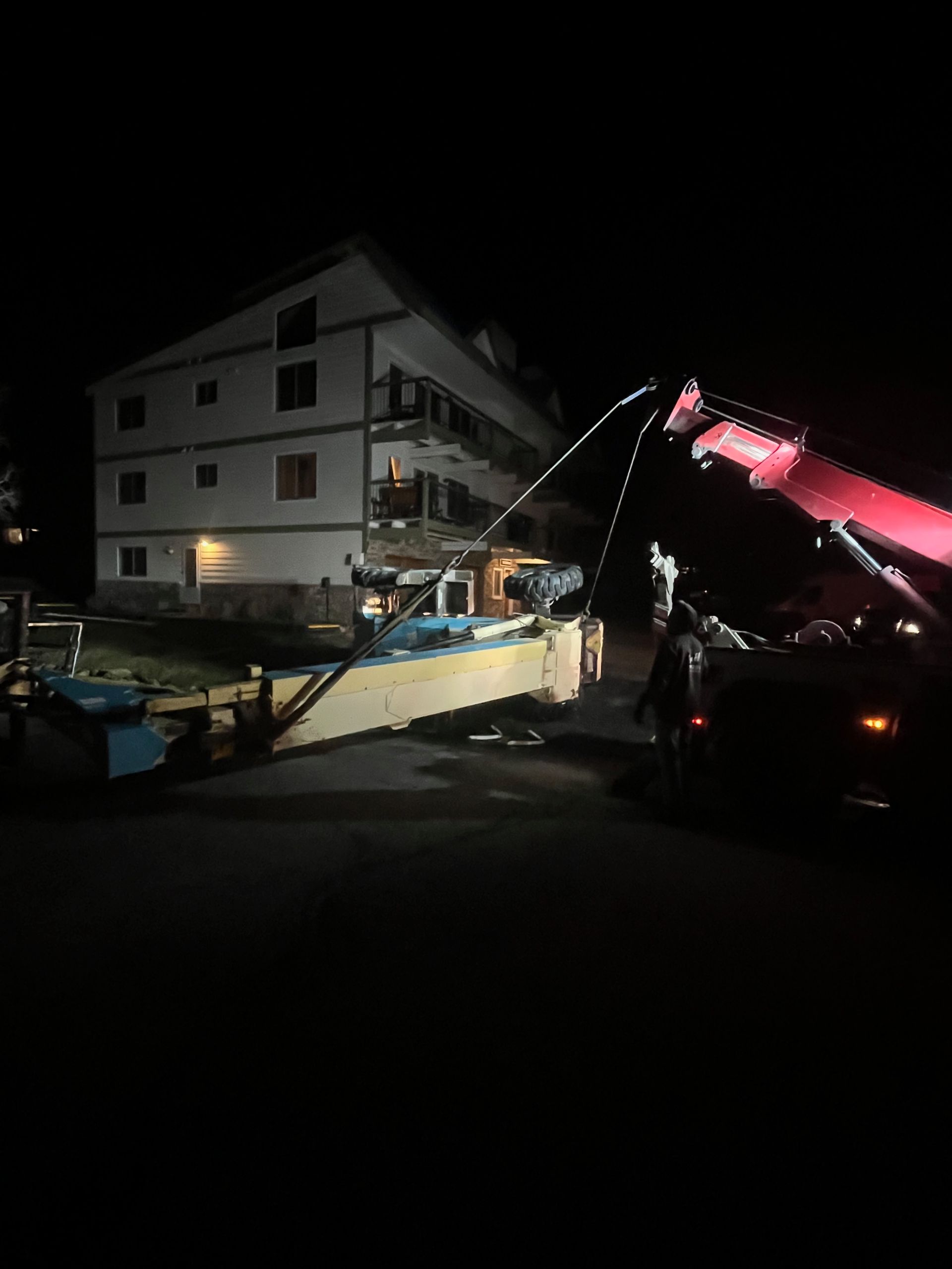 Crane lifting a long wooden structure near a multi-story building at night.