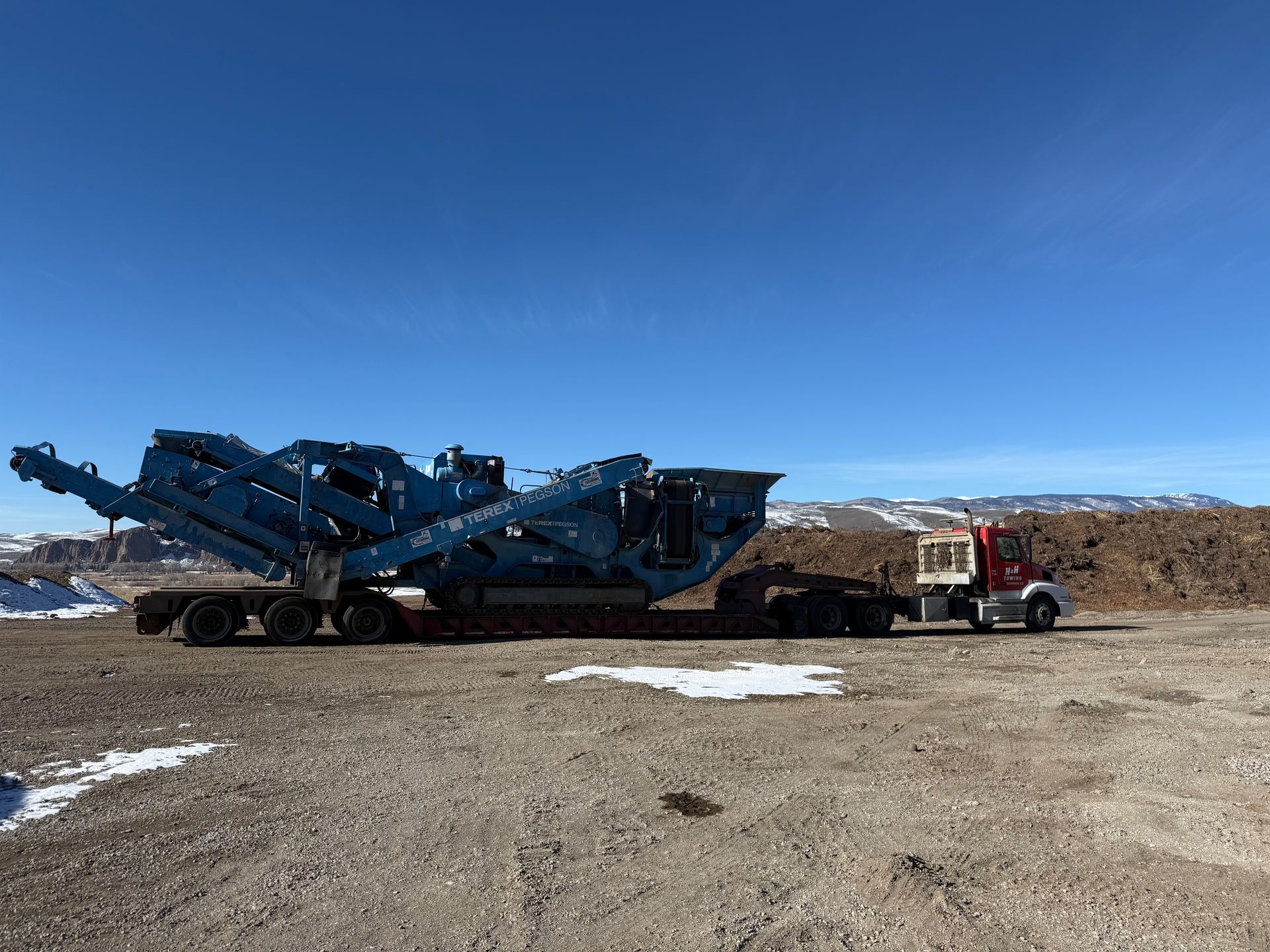 A blue industrial machine on a trailer is towed by a red semi-truck on a rocky surface, under a blue sky.