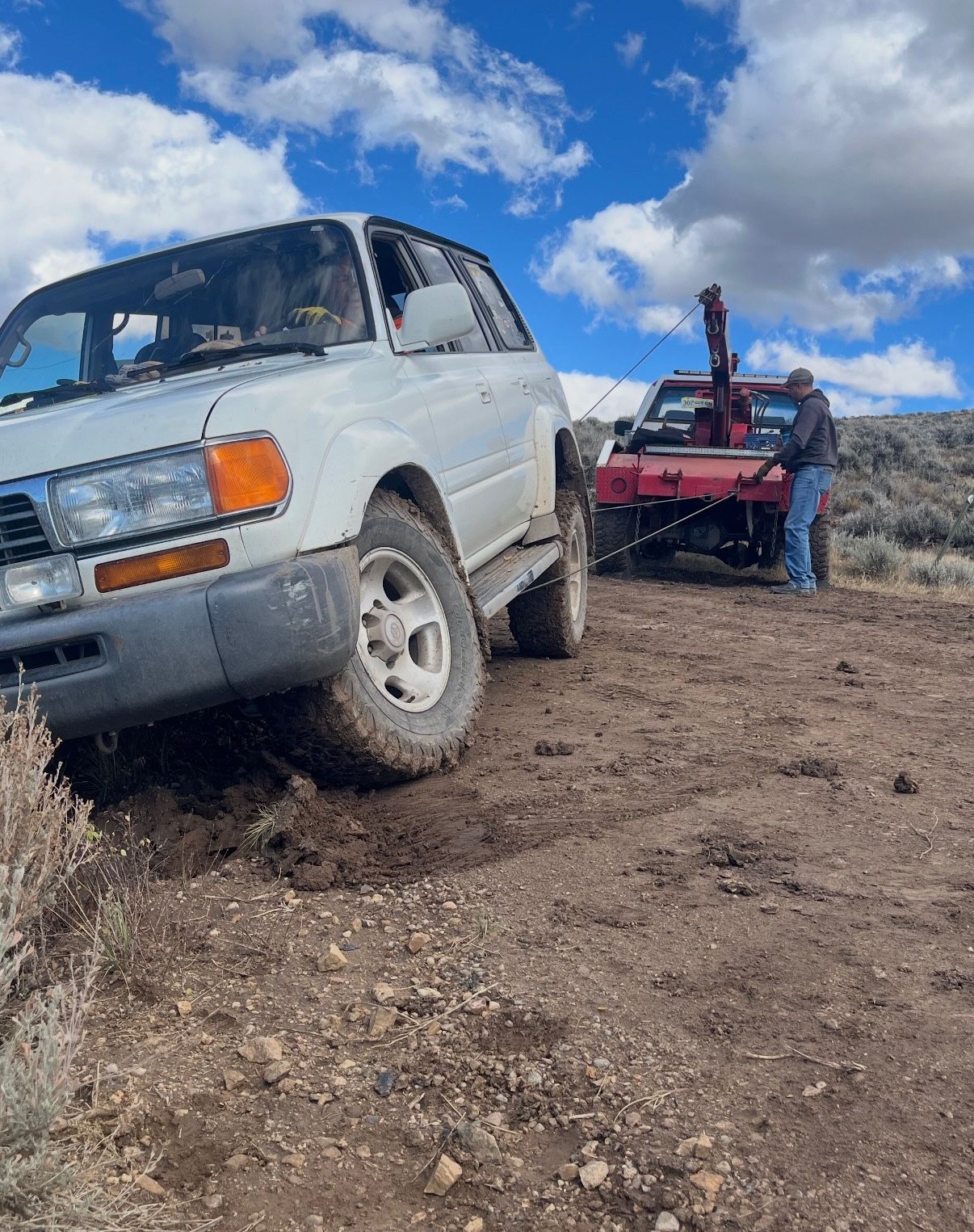 White SUV being towed out of dirt by red tow truck on a dirt road, under a cloudy sky.