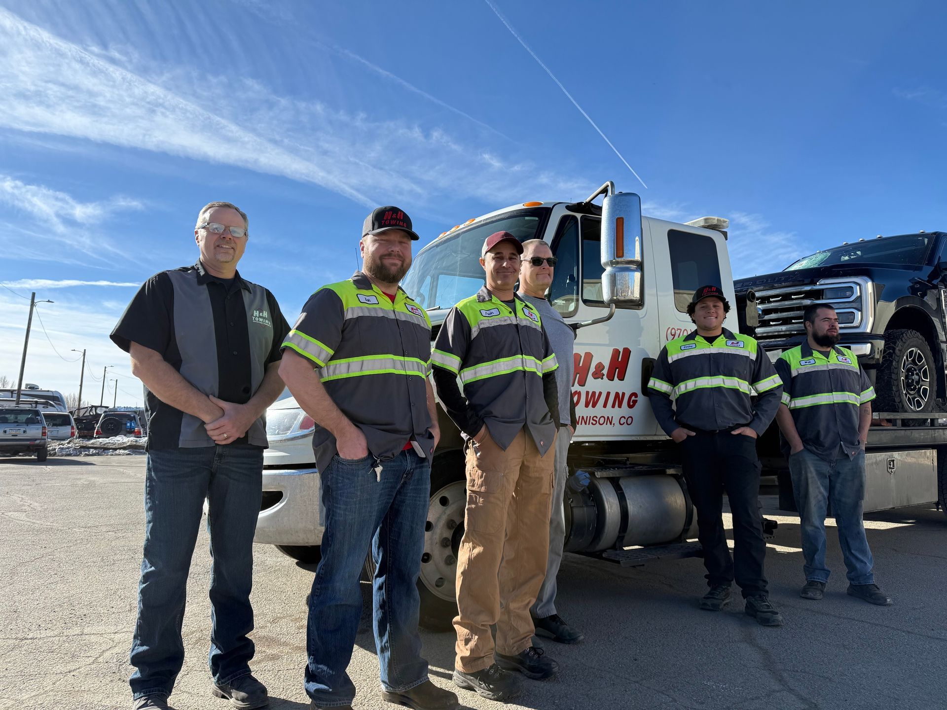 Six people in uniform stand near a tow truck. Sky is blue with few clouds. The truck has a black car on the platform.
