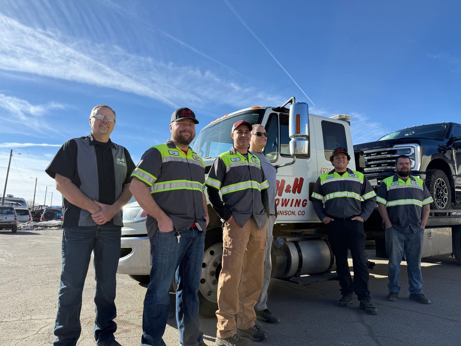 Six tow truck employees stand in front of a tow truck with a truck on its bed on a clear day.
