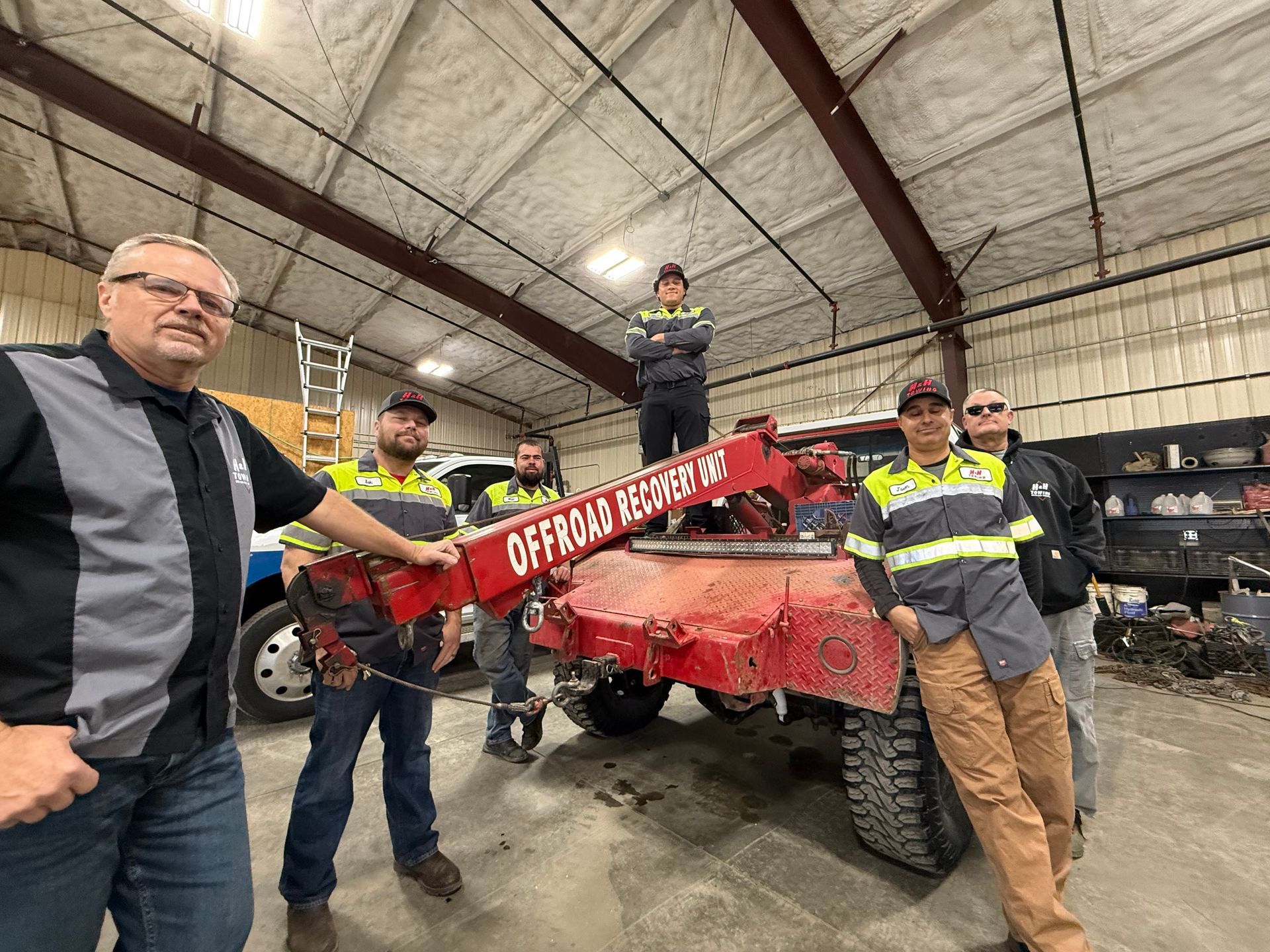 Group of men posing with a damaged tow truck inside a garage; one man is on the truck.