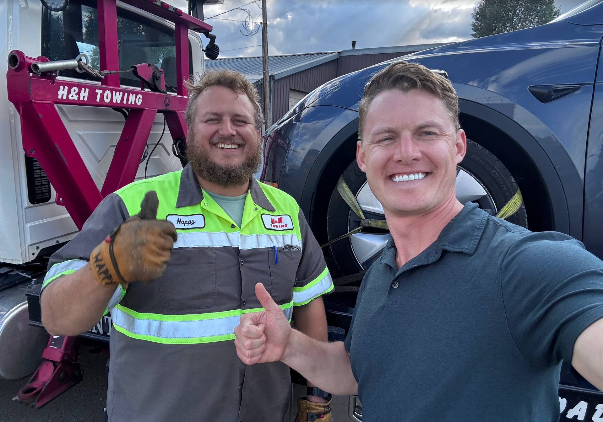 Two men with a tow truck smile, give thumbs up next to a car. One man wears a reflective vest. H&M Towing truck visible.