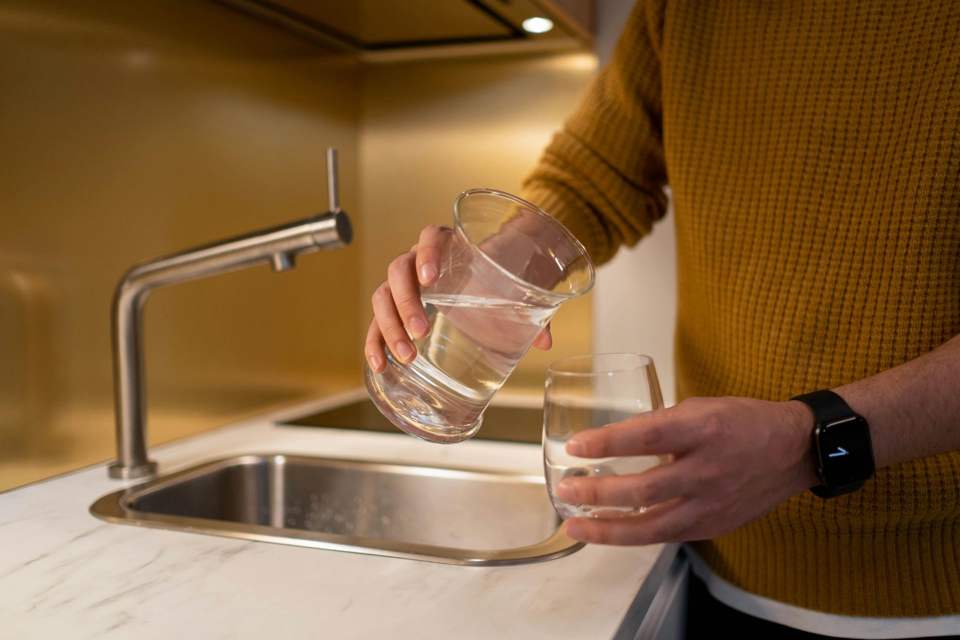 A person in a yellow sweater pours water from a glass pitcher into a glass over a kitchen sink.