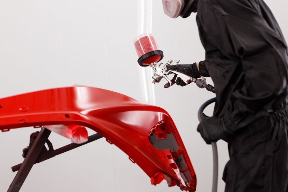 A technician in protective gear uses a spray gun to apply bright red paint to a vehicle bumper.