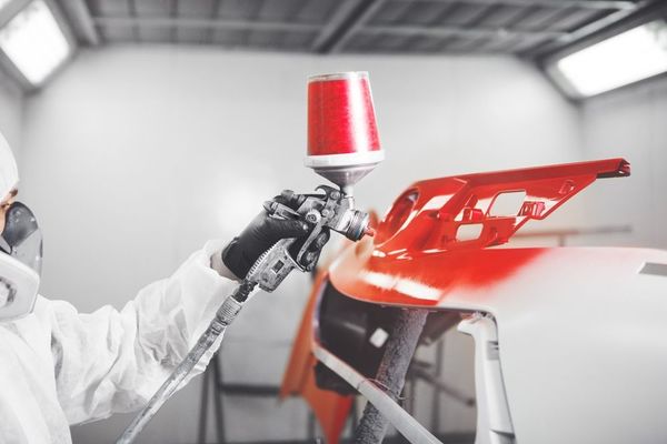 A worker wearing protective gear uses a spray gun to apply bright red paint to a car bumper in a workshop.
