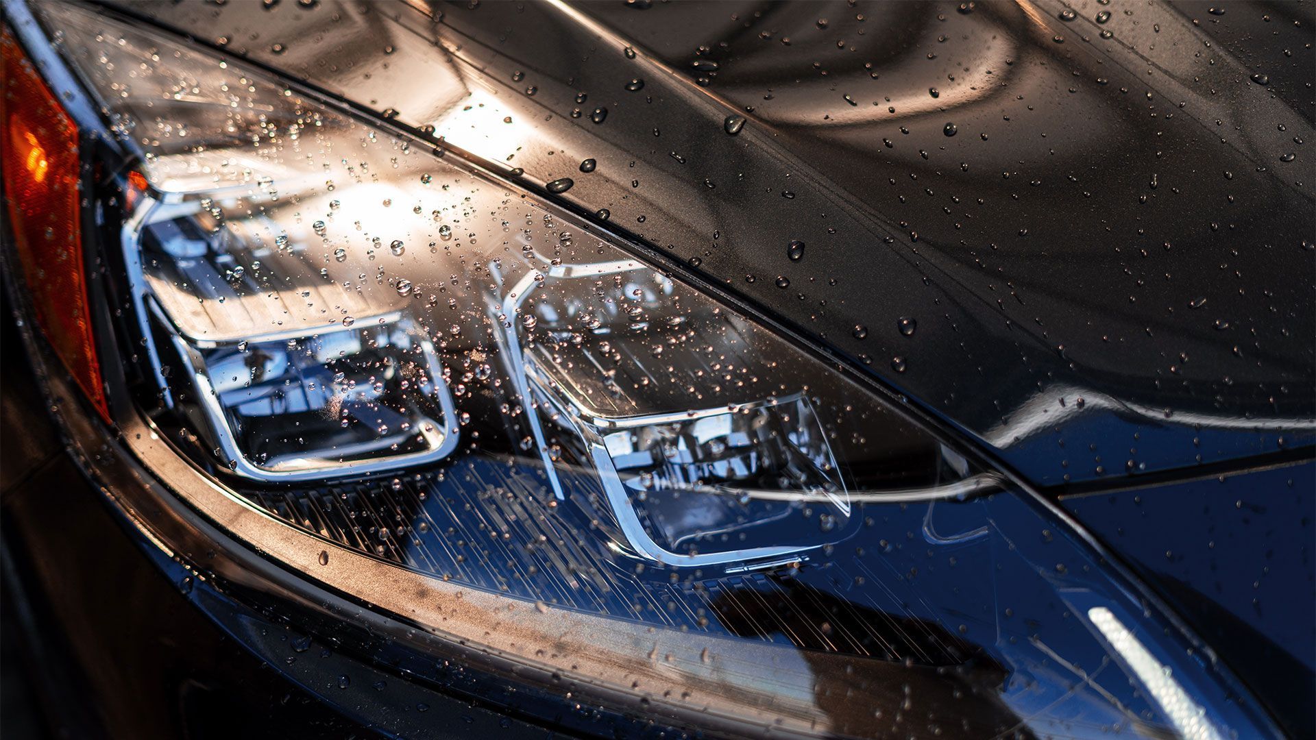 Close-up of a modern car's sleek black headlight assembly covered in raindrops, showing internal lighting components.