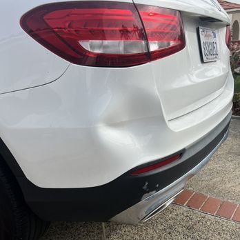 A white vehicle with a noticeable dent in the rear bumper and some paint damage on the lower black trim.