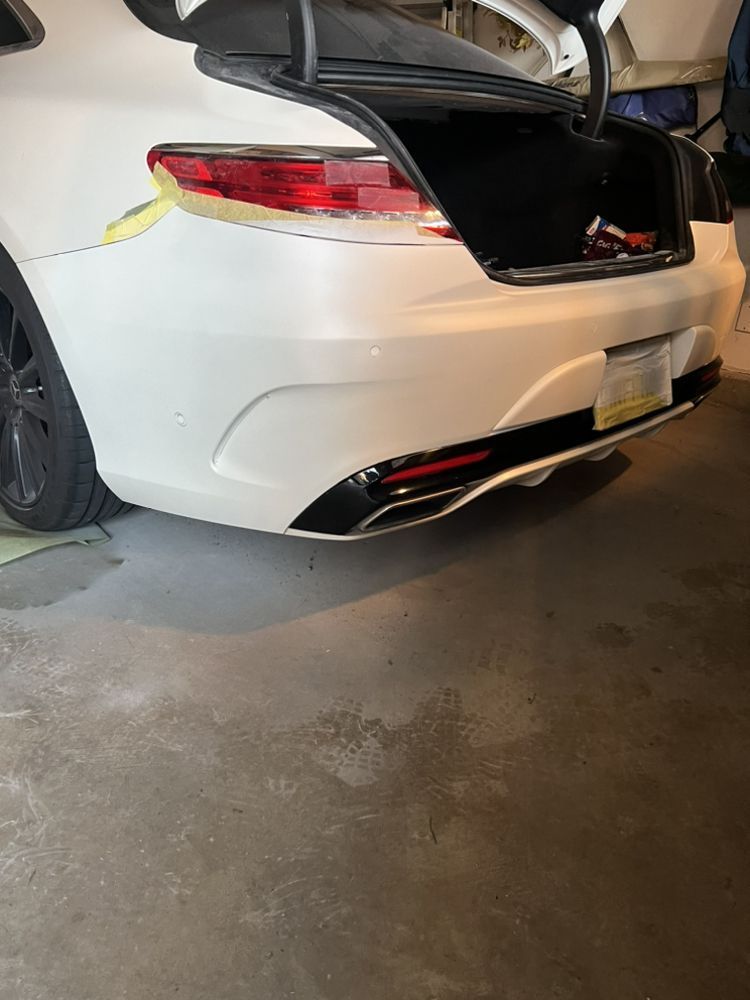 A white car parked in a garage with the trunk open and masking tape on the rear body panel near the taillight.