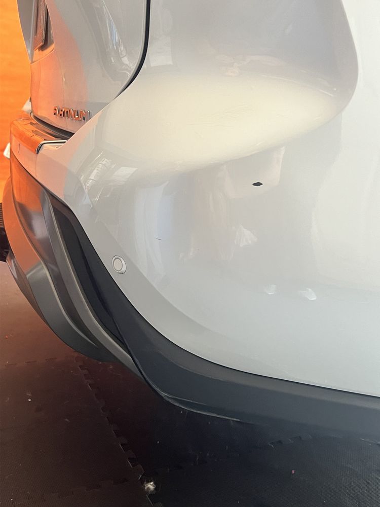 Close-up of a white car's rear bumper showing a circular parking sensor and a small black scuff mark.