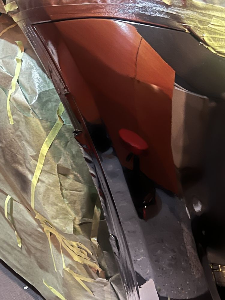 A close-up of a car panel being prepped for paint, showing red primer, masking tape, and protective paper covering.