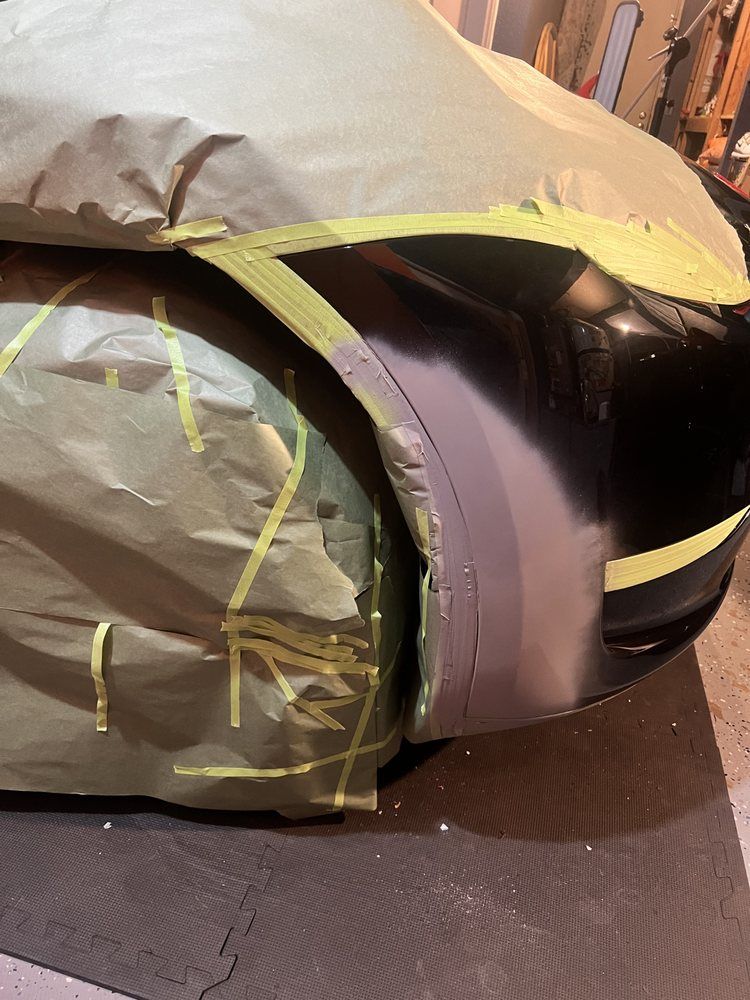 Car bodywork masked with paper and tape, showing a sanded, gray-primed fender section ready for painting.