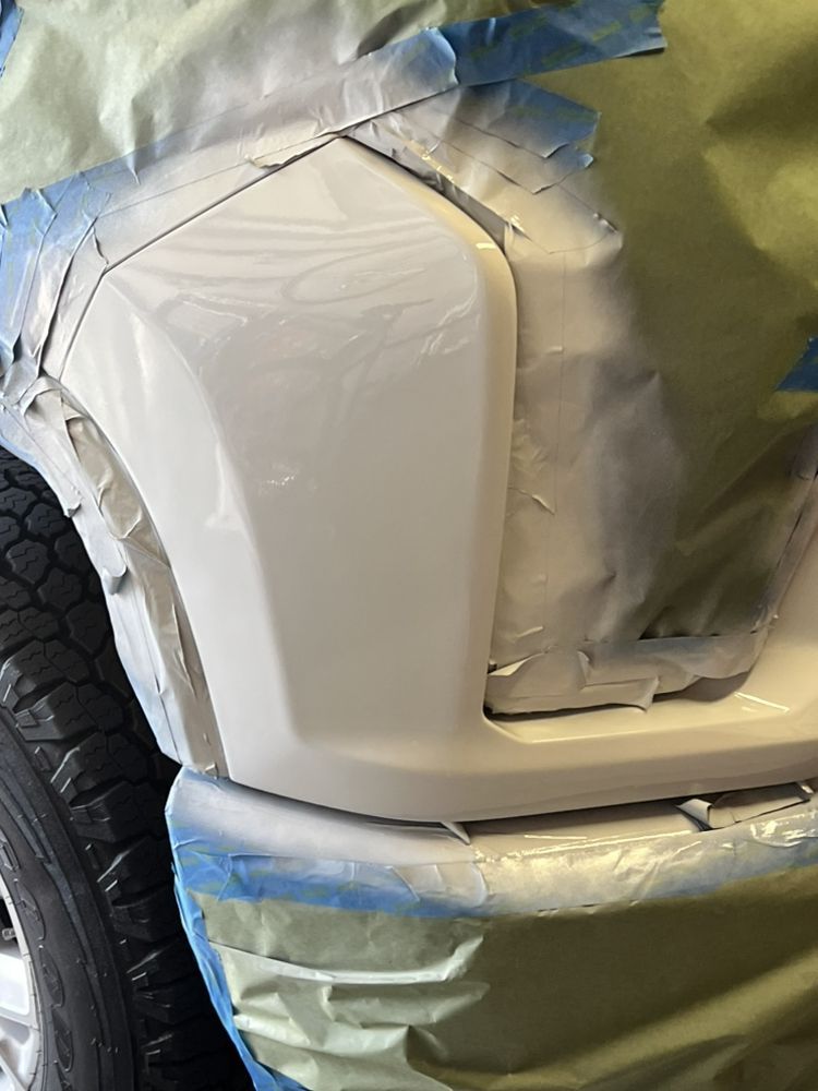 A vehicle's front panel, partially repainted in white, with surrounding areas masked off with blue tape and paper.