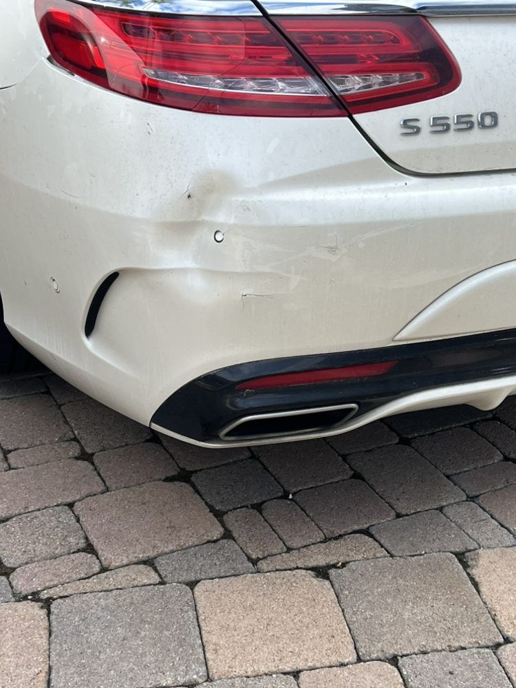 A dented white rear bumper on a Mercedes S550 parked on a paved driveway.