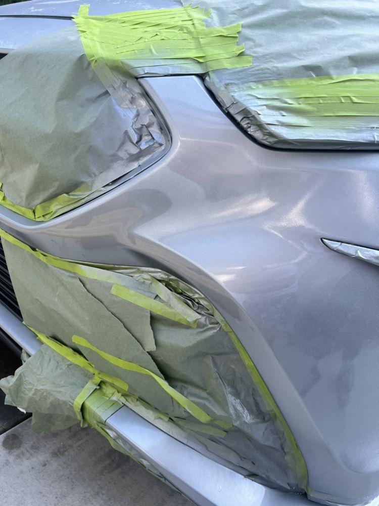 A silver car bumper with parts covered in green masking tape and paper in preparation for body repair or paint.
