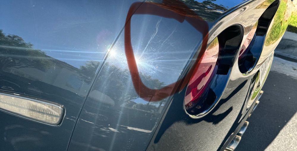 A close-up view of a dark car's rear panel showing a star-shaped crack in the paint near the taillight assembly.