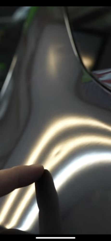 A finger points to a shallow, circular dent on a car's metallic body panel, highlighted by reflected light lines.