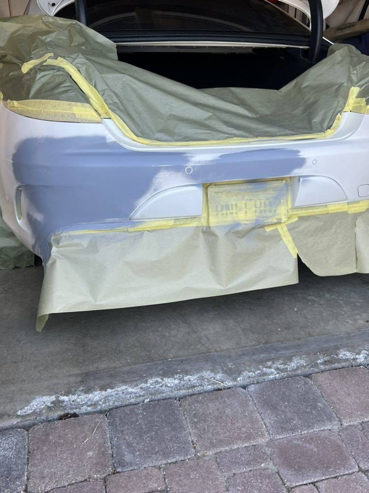 A car bumper prepared for painting with gray primer, masking tape, and protective paper covering the surrounding areas.