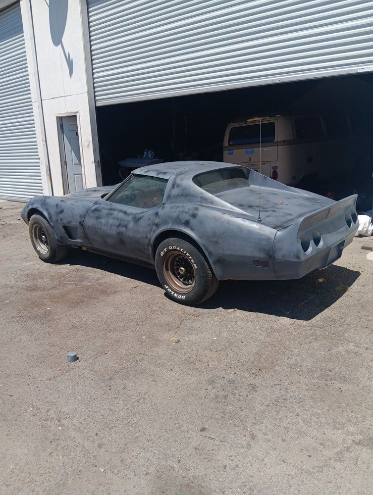 A dark grey, project-condition Chevrolet Corvette C3 parked outside a garage on a sunny day.
