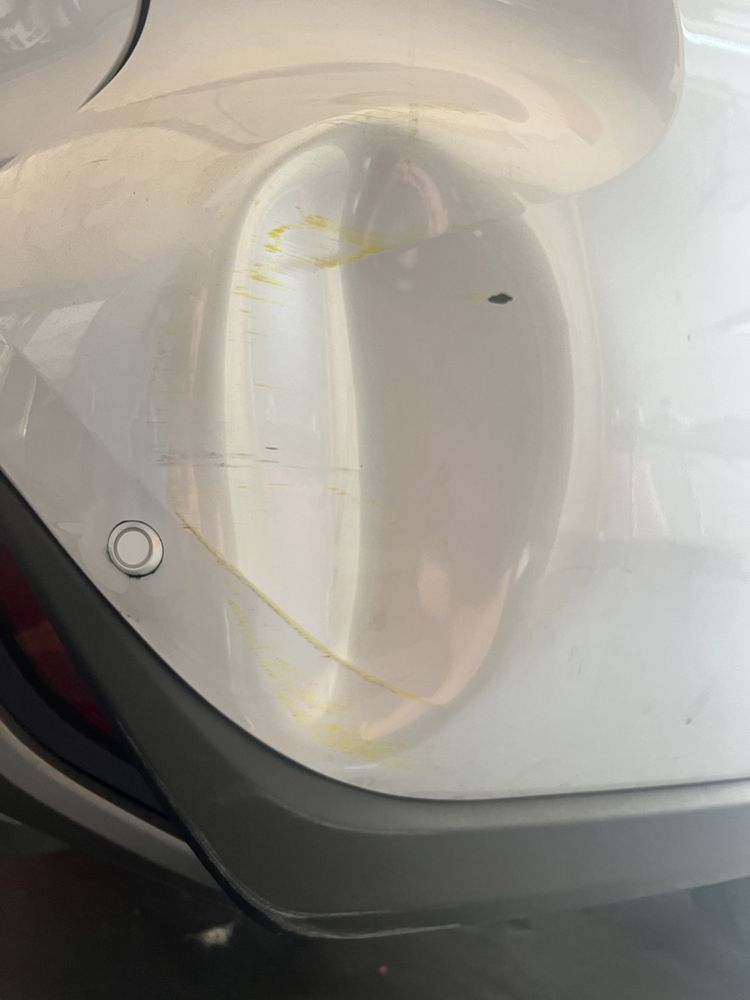 A close-up of a white car bumper featuring a dent and yellow paint transfer scuffs near a parking sensor.