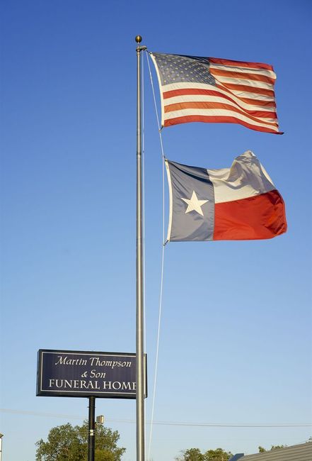 Two flags waving on tall poles: the U.S. flag and the Texas state flag above a roadside sign.