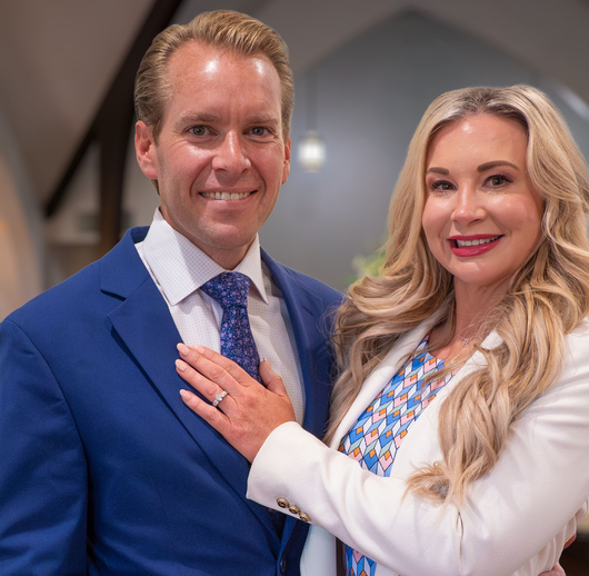 Two smiling people posing indoors, woman resting hand on man’s chest, both in business attire