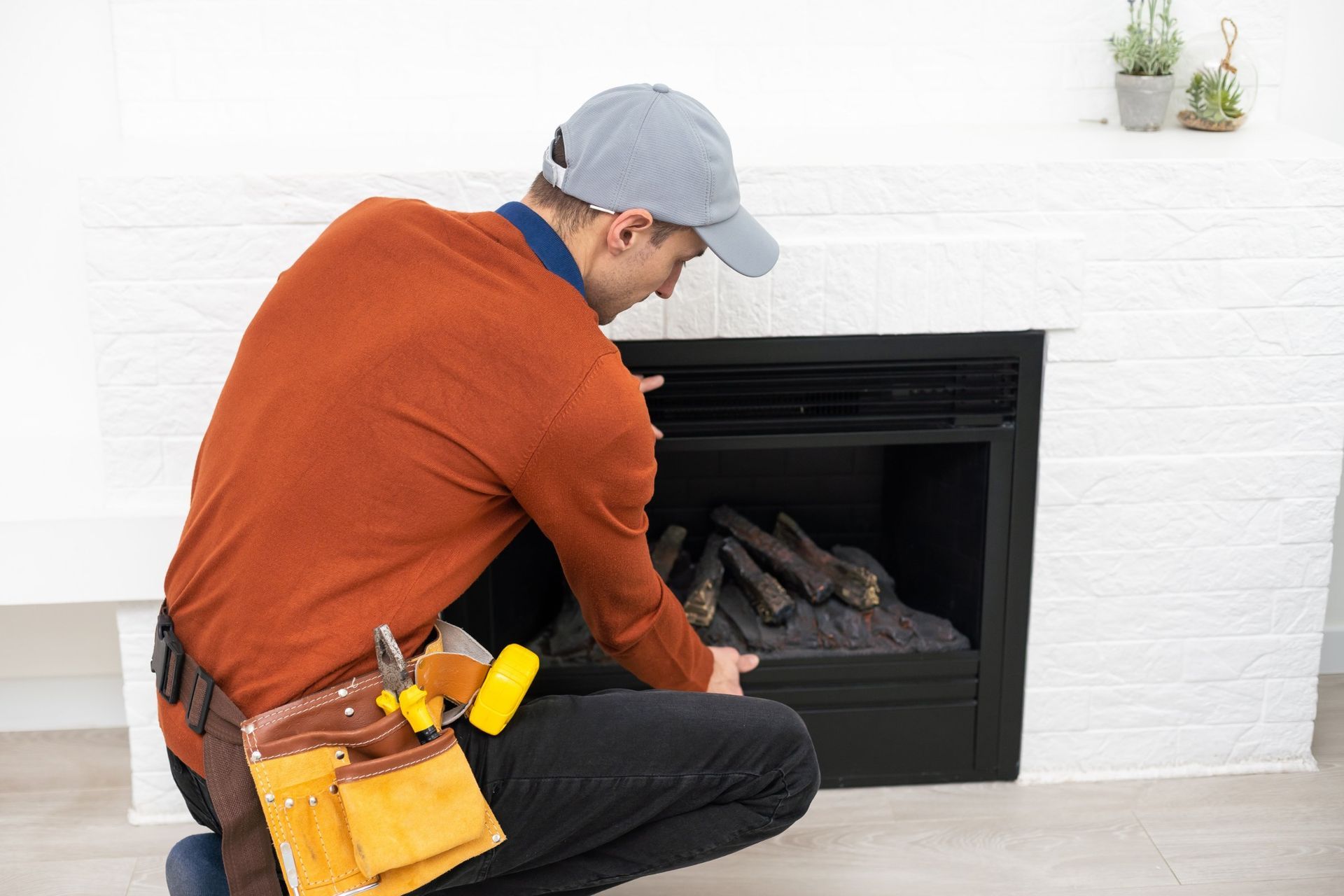 Man in orange shirt and tool belt examining a black fireplace. White brick wall background.