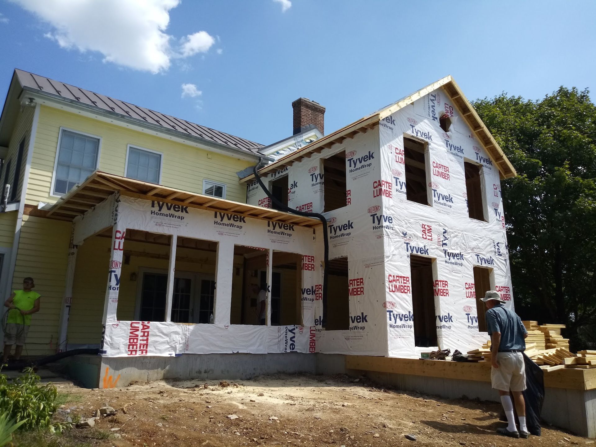 House under construction; workers on site, wrapped in Tyvek. Yellow house connected to new frame. Sunny day.