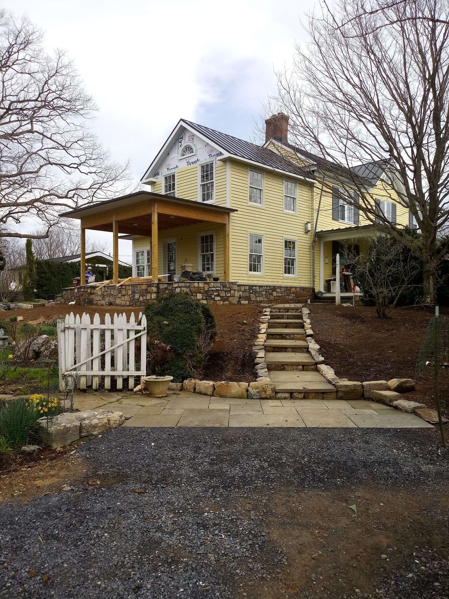 Yellow farmhouse with porch, steps, and white picket fence on a hill.