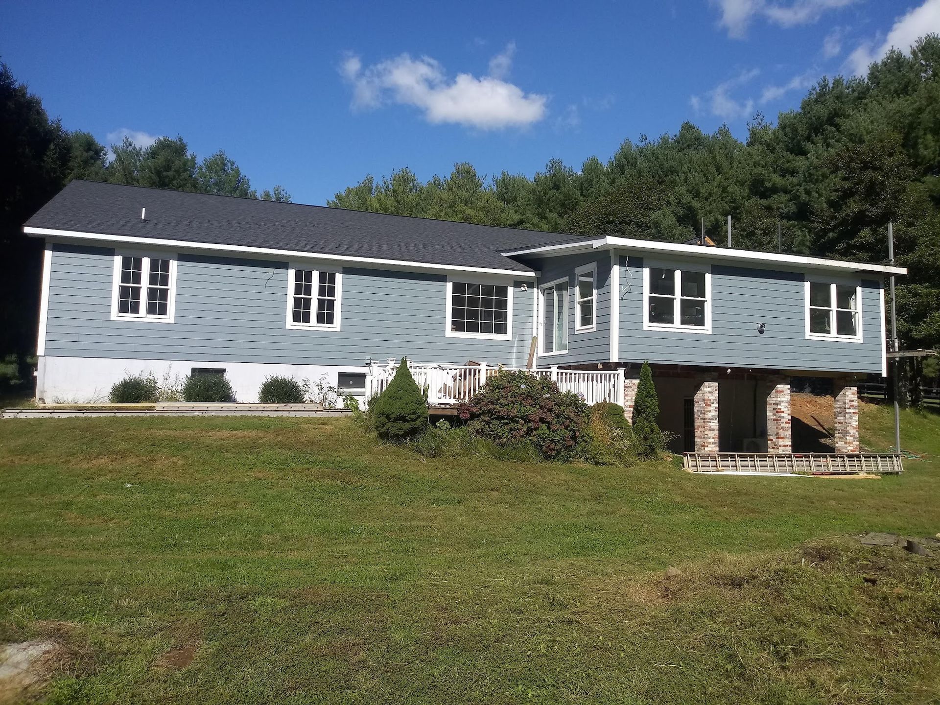 Blue-sided house with white window frames and a gray roof, set on a green grassy hill, with a forest in the background.