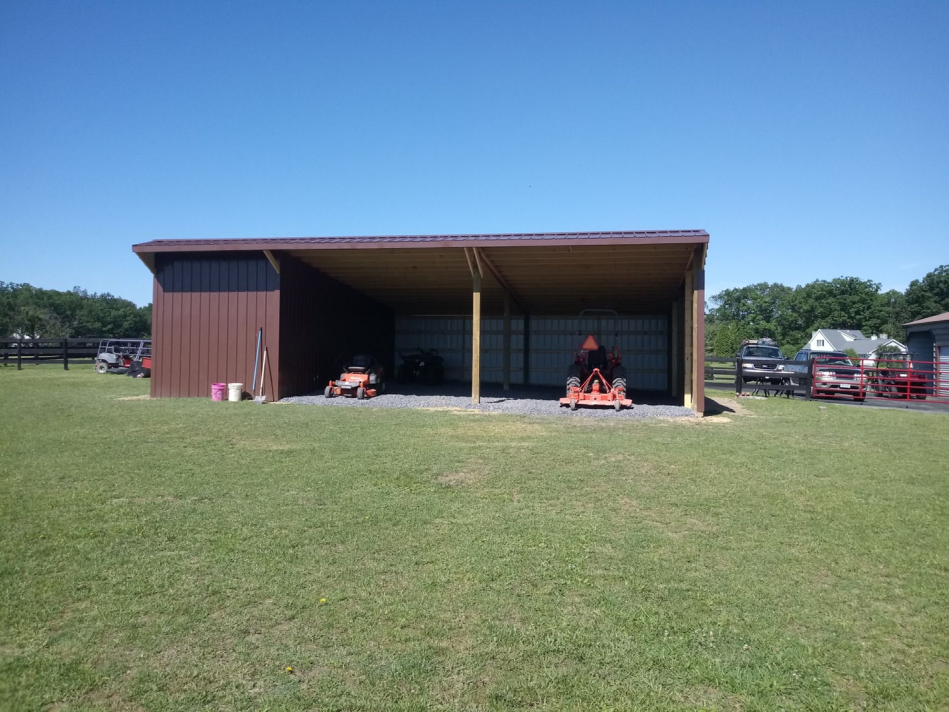 Brown open-sided metal shed in a grassy field, housing two lawn mowers. Blue sky overhead.