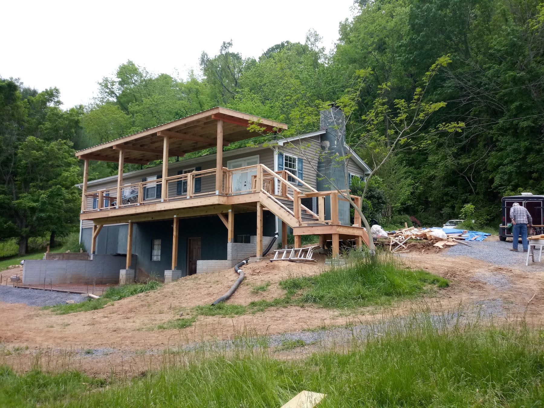 A house with a deck under construction, surrounded by trees and a man working nearby.