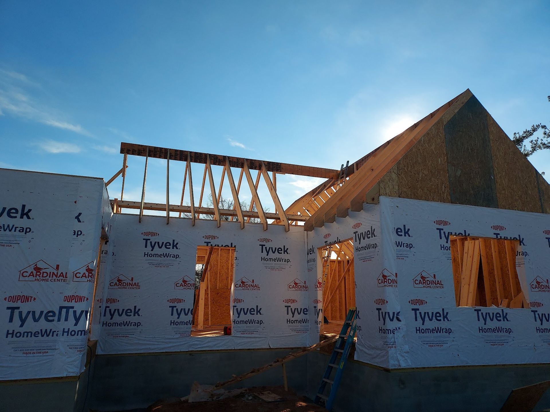 House under construction, frame and roof visible, wrapped in Tyvek, under a blue sky.