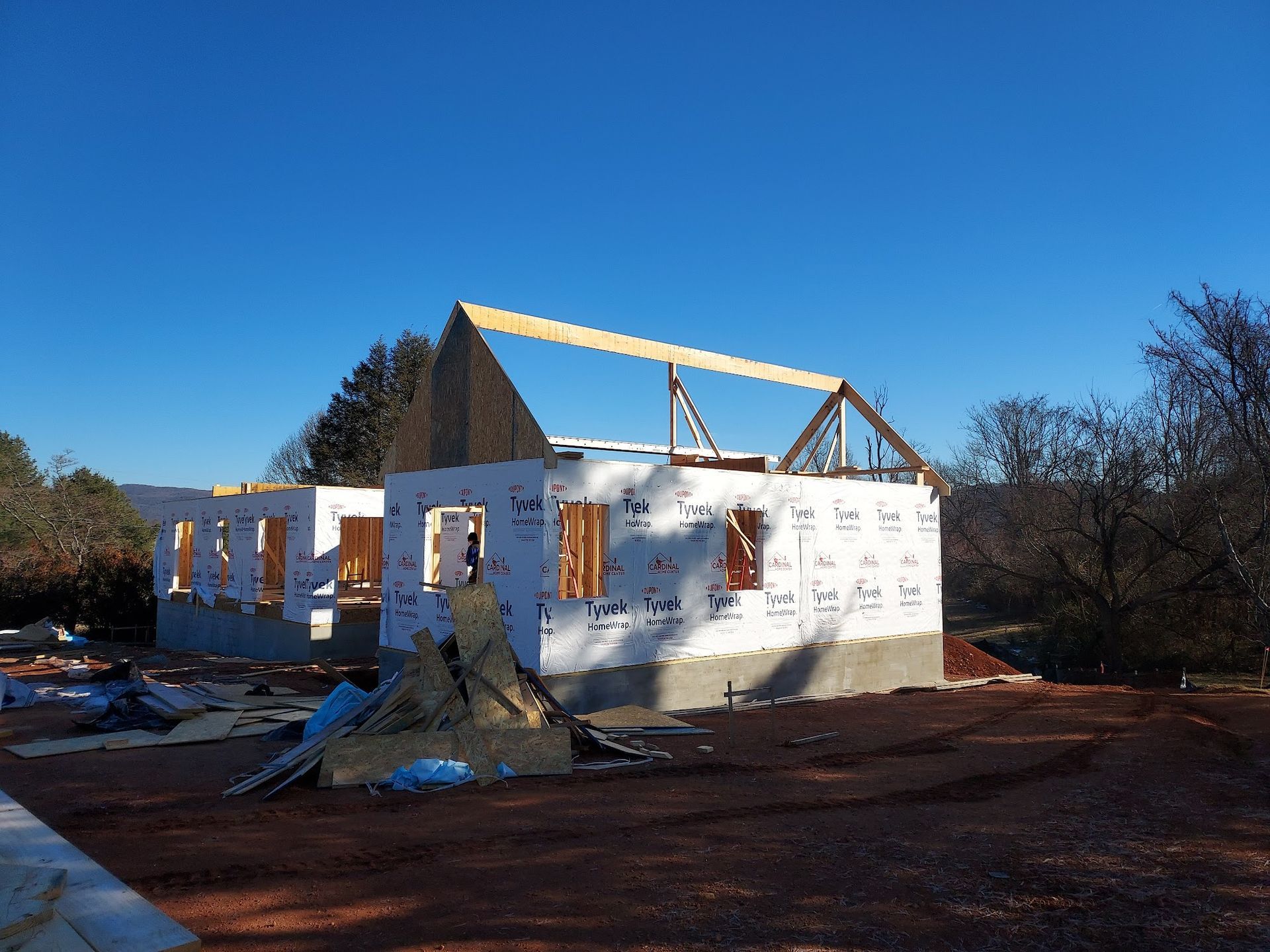 Construction site of a house; wood frame, roof trusses, blue sky, and protective wrap are visible.