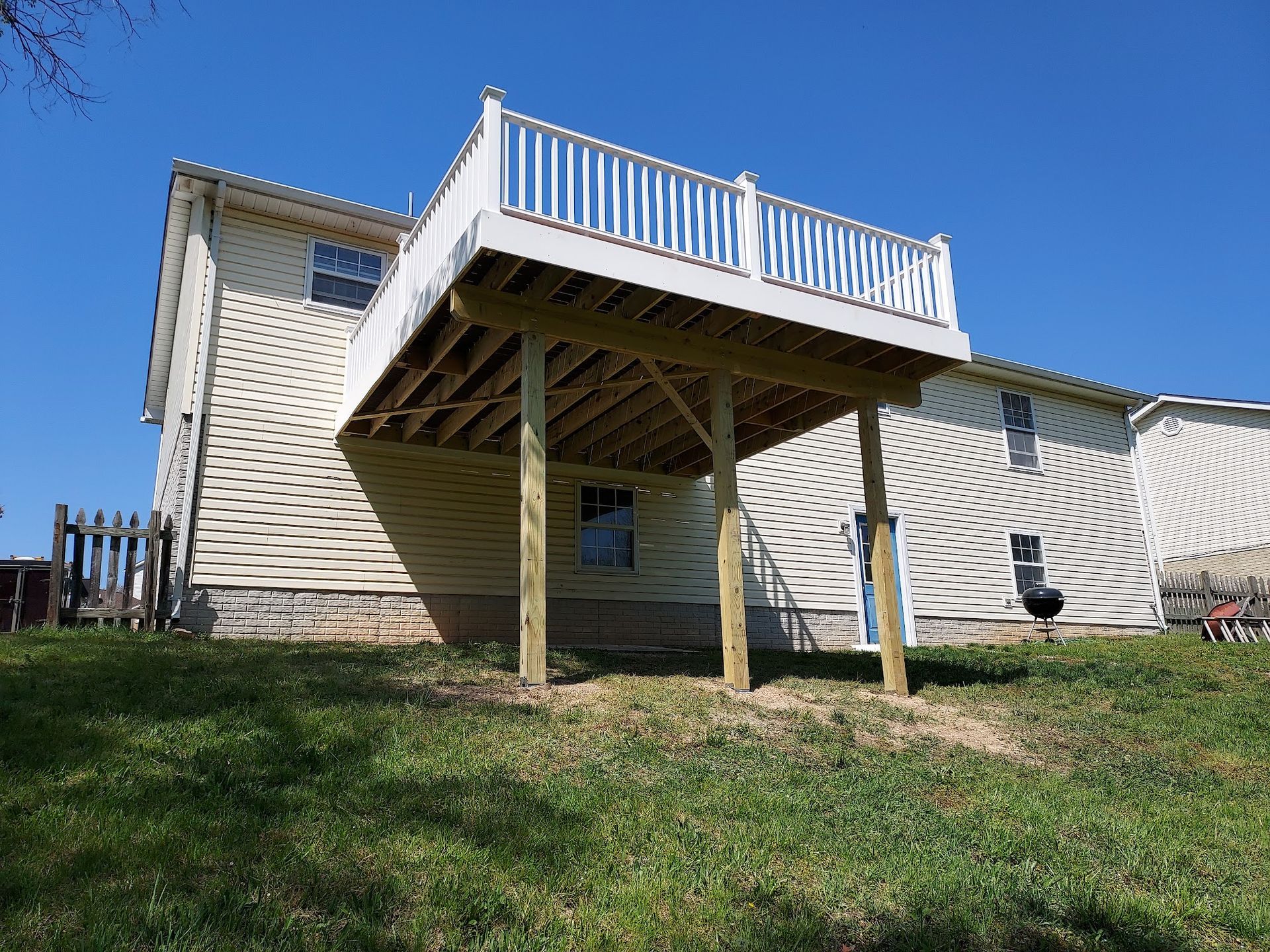 Raised wooden deck attached to a light-yellow house, with white railing. Green grass in foreground, blue sky background.