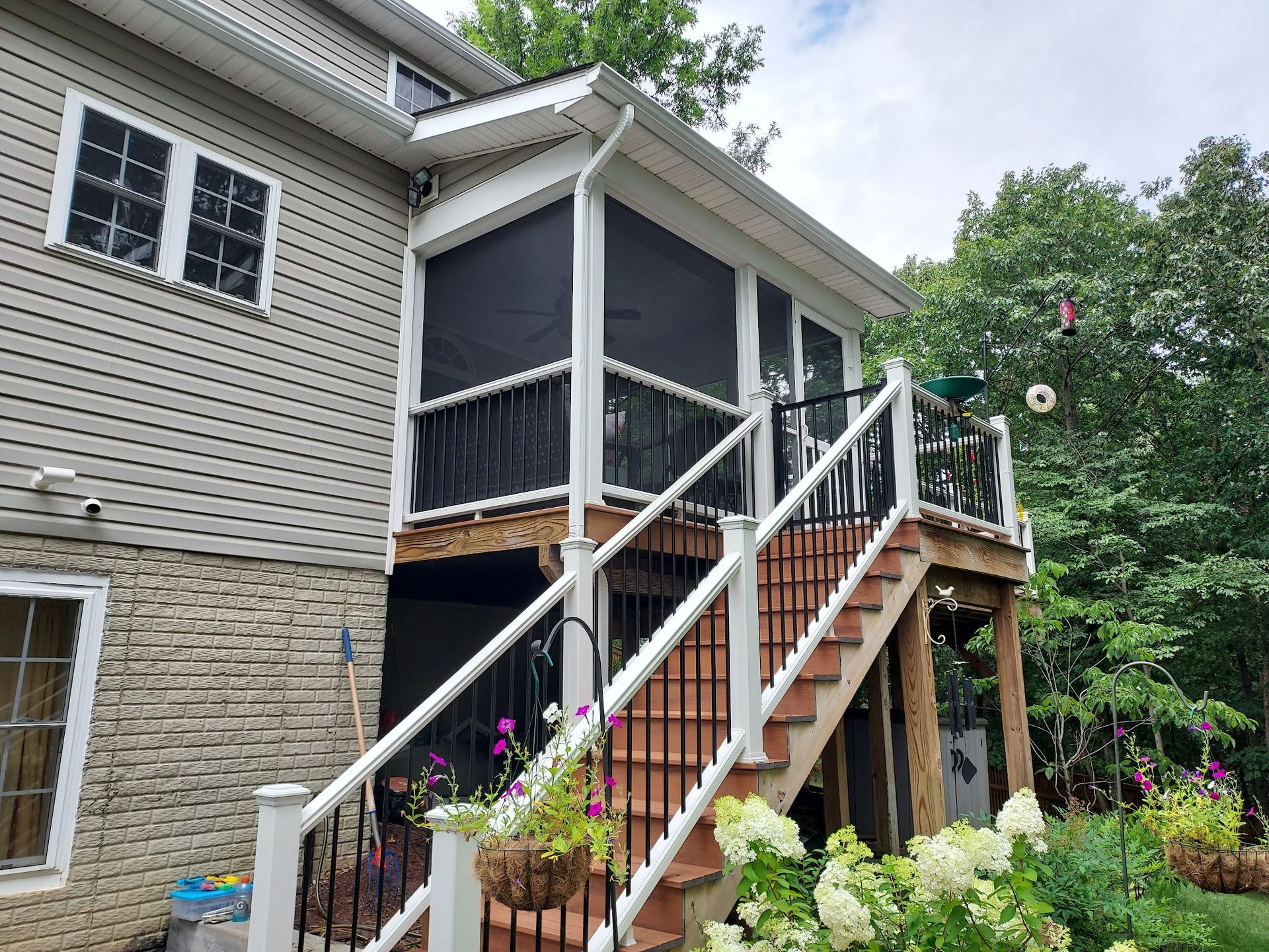 Screened porch and deck with stairs attached to a beige sided house; surrounded by trees and flowers.