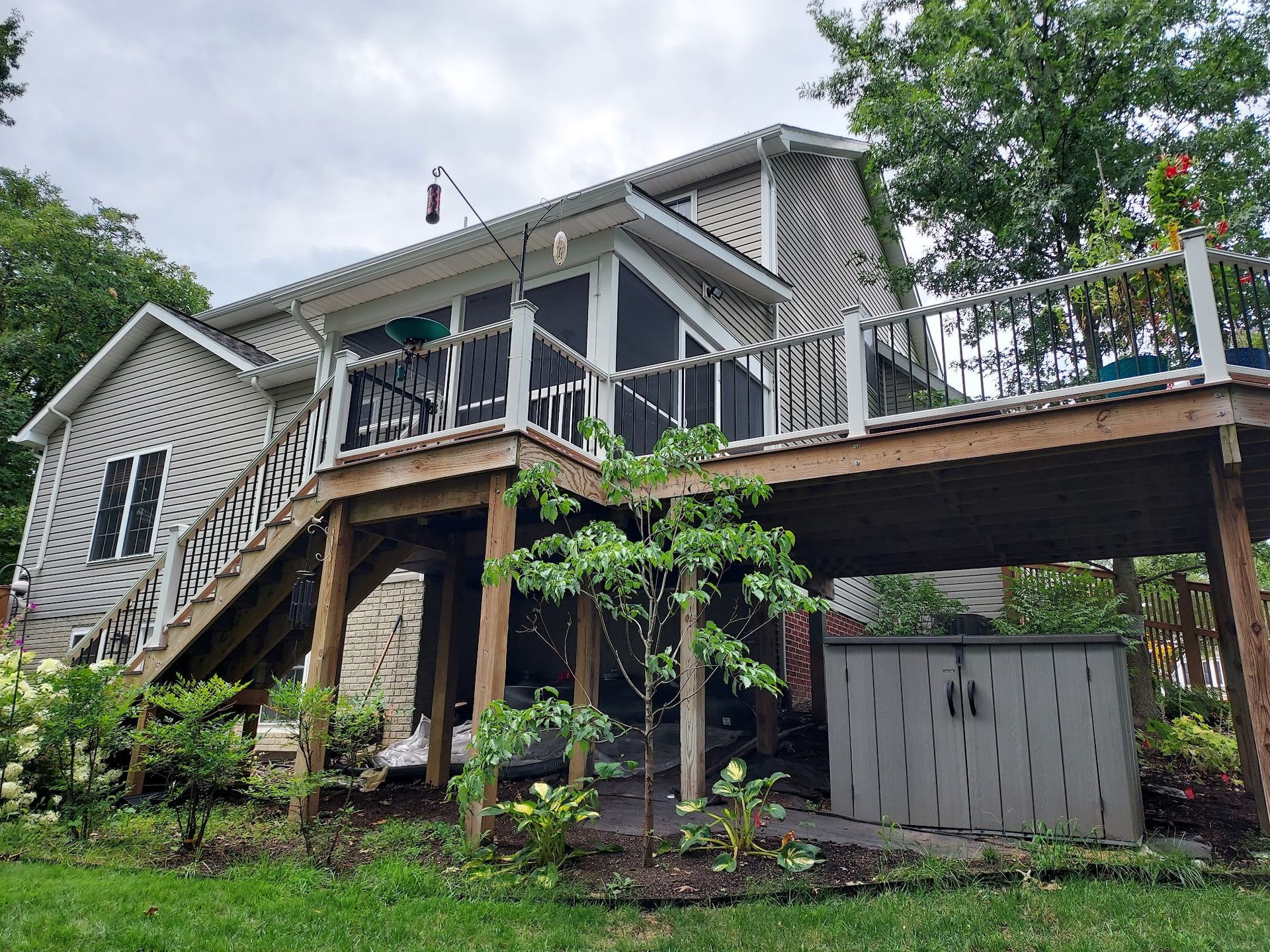 Wooden deck with screened-in porch, overlooking yard and shed. House with gray siding and lush greenery.