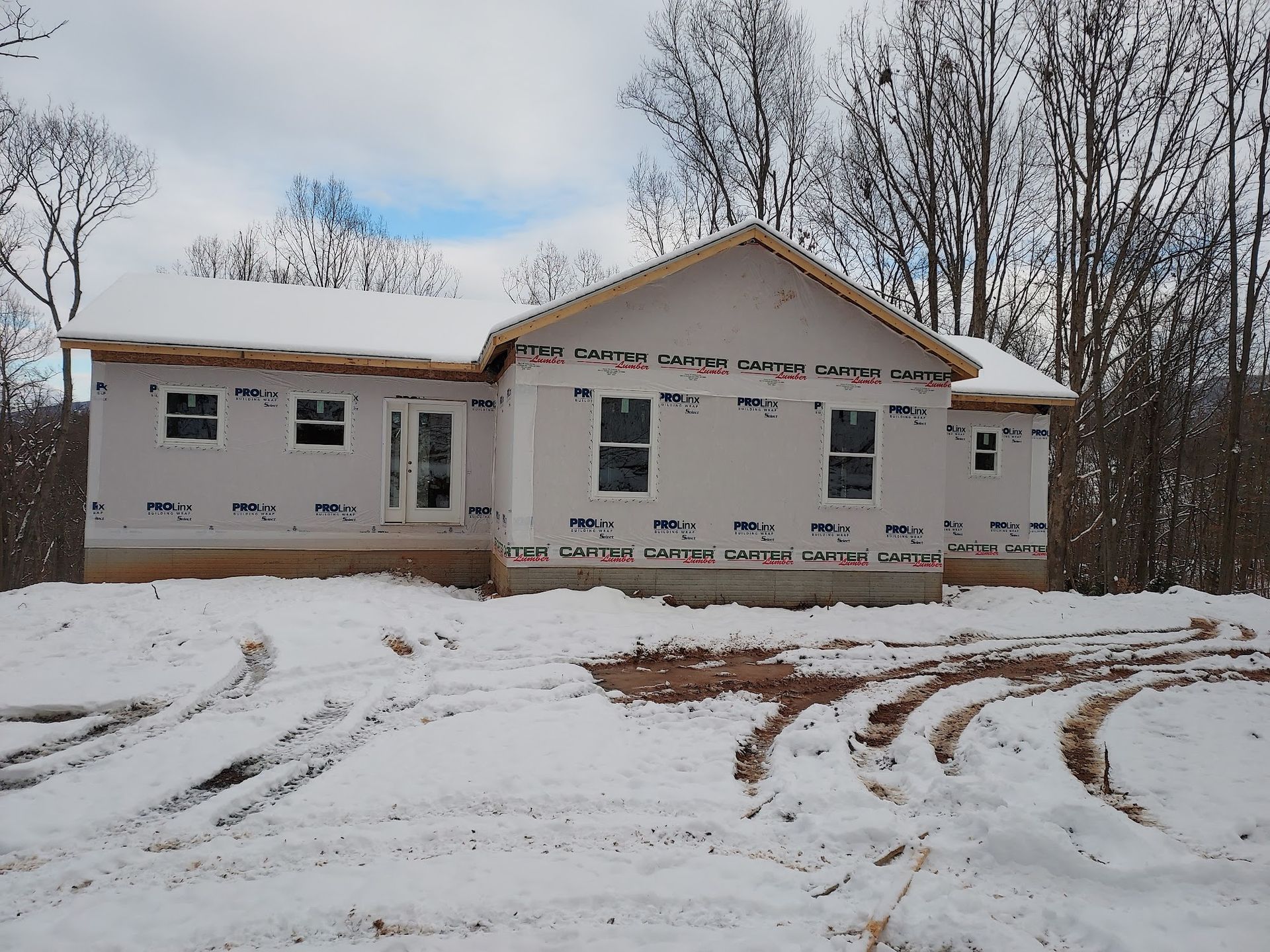 House under construction, covered in blue sheathing, in snowy setting with tire tracks.
