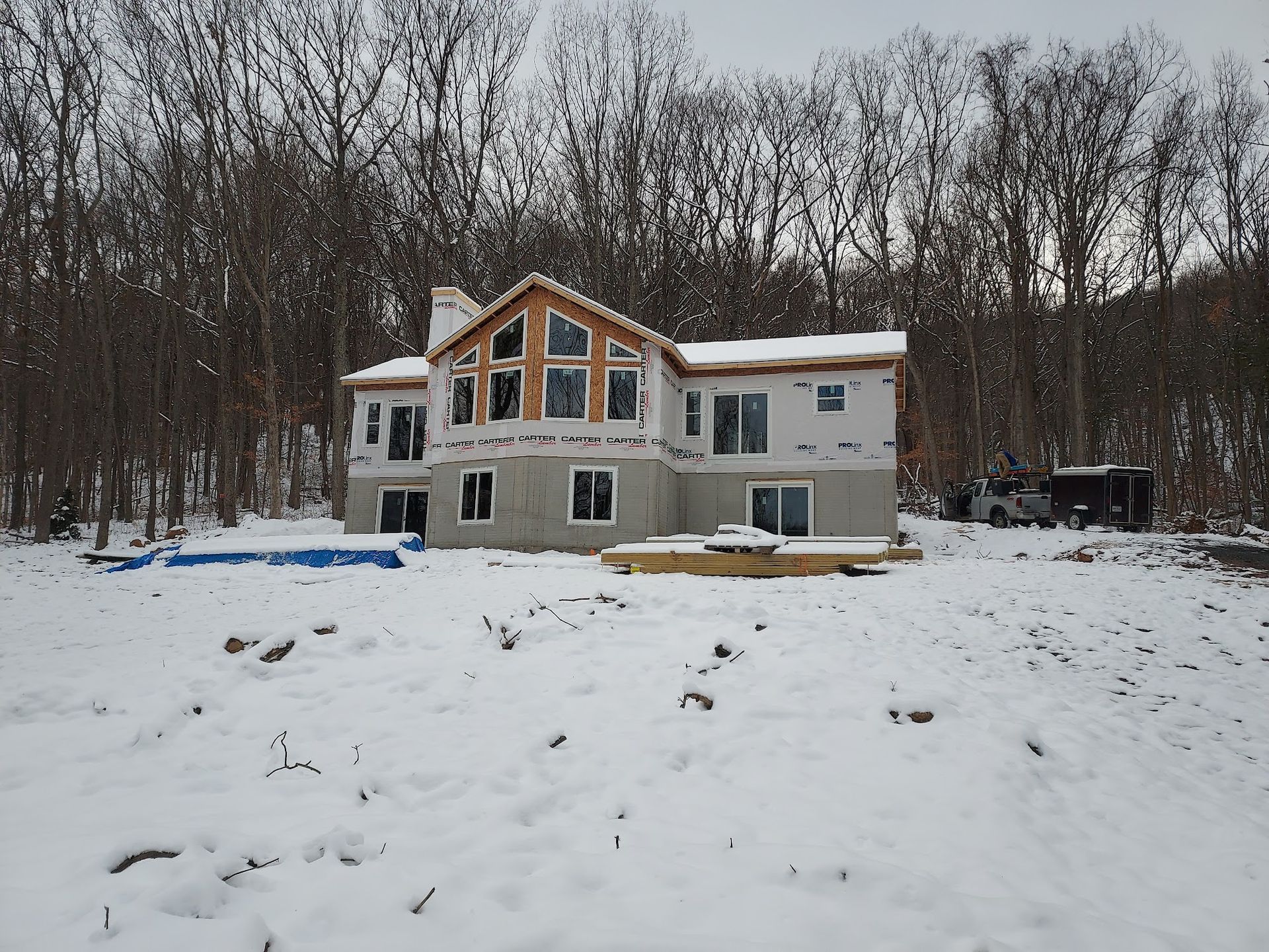 Two-story house under construction in a snowy forest. Unfinished exterior with large windows and a deck.