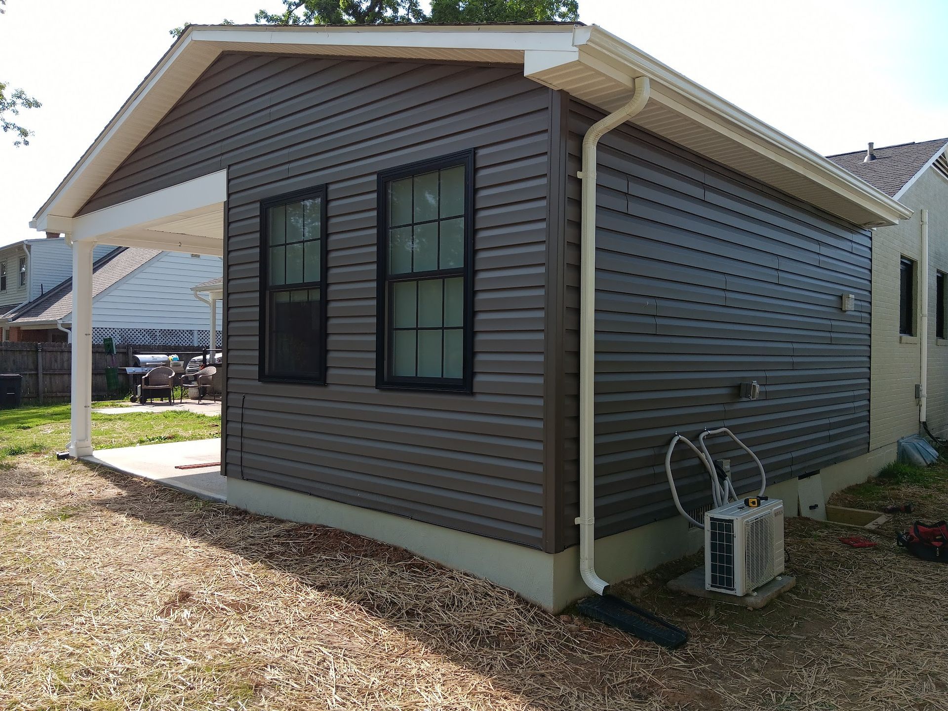 Brown-sided shed with white trim, a porch, two windows, and an air conditioning unit.